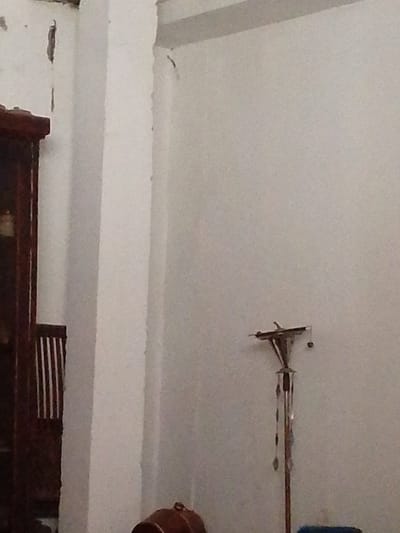 This is a close-up, slightly blurry, indoor photograph taken at Atamo Sur, Venezuela. The scene is dominated by a large, off-white wall that occupies most of the frame. To the left, a dark wooden cabinet or furniture piece is visible, with vertical slats in its lower section. A white column or structural element runs vertically down the center-left of the image. The wall shows some signs of wear and tear, with a few dark, elongated cracks or marks near the top and along the column.
In the mid-right of the image, a decorative item resembling a wind chime or mobile hangs on the wall. It has a central pole with several delicate, metallic elements hanging from it, catching the light. A small, dark sphere is also attached to it. The lighting suggests it's daytime, but the interior setting is dimly lit, contributing to the overall grainy quality of the image. No people are visible. The focus is on the interior details of the room, hinting at a lived-in but perhaps somewhat neglected space. No text is visible in the image.