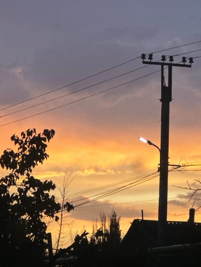 This is a photograph taken at dusk in the city of Novopokrovka, Kyrgyzstan. The sky is a gradient of colors, transitioning from a deep purple at the top to a vibrant orange and yellow near the horizon, indicating the sun is setting. Clouds are present in the sky, illuminated by the fading sunlight. A prominent telephone pole with insulators and wires runs across the frame, silhouetted against the colorful sky. A street lamp attached to the pole is illuminated. In the foreground and to the left, the dark silhouette of a leafy tree stands out. Further to the right, the dark silhouette of a building with a pitched roof is visible, along with other dark shapes that appear to be trees and structures. The overall scene depicts a twilight view of an urban or semi-urban area.