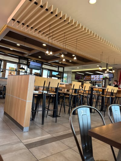 The image shows the interior of a fast-food restaurant, likely a Burger King, located at Mittelstraße 1 in Mülheim-Kärlich, Germany. The restaurant features a modern design with a slatted wood ceiling, pendant light fixtures, and a variety of seating options including high-top tables with bar stools and lower tables with chairs. The color palette consists of warm wood tones, black accents, and neutral flooring. A digital menu board displaying food items is visible in the background, along with a counter area where a male employee in a dark shirt is standing. The restaurant appears to be open for business, with some tables occupied by patrons.
