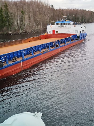 A long, flat-bottomed barge, identified as "ОКСКИЙ 10" (OKSKIY 10) in Cyrillic lettering, navigates a tranquil river or canal. The vessel features a white superstructure with a blue roof, a red hull, and a rust-colored deck, complemented by blue railings and supports. Orange life rings are visible on its upper deck, which houses a bridge/wheelhouse equipped with navigational equipment like radar and antennas. The barge is actively in motion, generating a visible wake behind its stern, as captured from the perspective of another vessel or structure partially visible in the bottom foreground.
The surrounding landscape consists of wooded banks dominated by bare deciduous trees, possibly birch, interspersed with some evergreen conifers, suggesting a setting in late autumn, winter, or early spring. The sky is overcast, casting a soft, diffused light over the scene. Draft marks, indicating measurements such as '40', '45', '50', '55', and '60', are clearly visible on the red hull near the waterline, providing an indication of the vessel's current depth in the water. This functional scene, depicting a working vessel on a waterway, is set in Uzhla, Russia, which aligns with the Cyrillic text displayed on the ship.