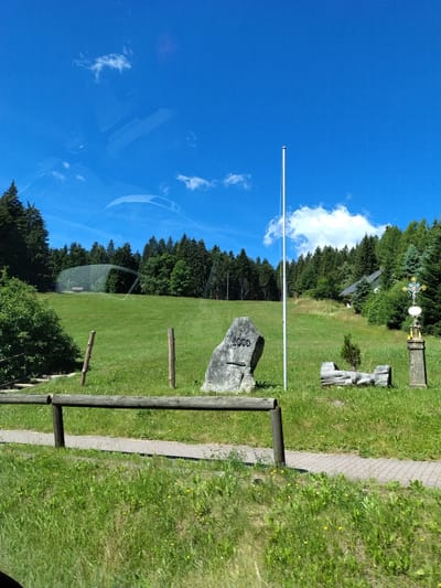 The image captures a picturesque outdoor scene in Kubova Huť, Czechia, on a bright, sunny day with a clear blue sky. The foreground features a paved walkway running horizontally across the lower portion of the frame, bordered by lush green grass dotted with small yellow wildflowers. A rustic wooden fence made of logs runs parallel to the walkway, separating it from the grassy slope beyond.
On the grassy slope, a large, rough-hewn stone stands prominently, inscribed with the number "1000". To the right of the stone, a tall, thin white flagpole is planted. Further to the right, a decorative stone monument with a cross sits on a pedestal. Behind these elements, a gentle hill rises, covered in vibrant green grass and a dense forest of coniferous trees. A small building with a dark roof is partially visible nestled within the trees on the hillside. The overall impression is one of natural beauty and tranquility, possibly a park or a scenic overlook. There are no people visible in the image.