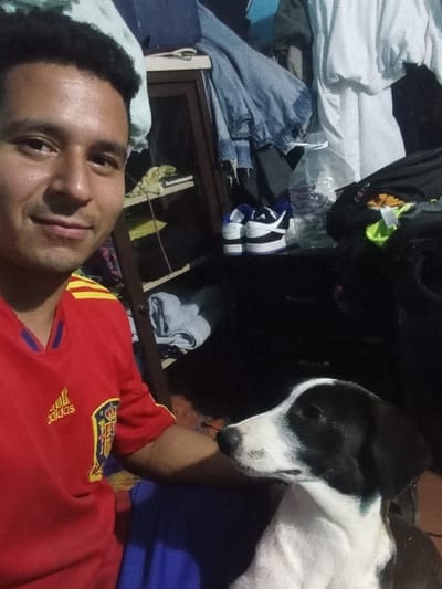 The image displays an indoor scene in Cujisal, Venezuela, featuring one male human subject and one canine subject. The male, positioned on the left, is an adult with dark, short, curly hair and a light complexion. He wears a red long-sleeved athletic shirt with yellow accents, an Adidas logo, and a crest resembling that of the Spanish national football team. He is looking directly at the camera with a neutral to slightly smiling facial expression. To his right, a black and white dog is visible, with black patches on its head and ears, and a white snout, forehead, and body. The dog is oriented to its right, away from the camera. The background depicts a residential interior with various objects. On the left, a dark wooden shelf unit holds unspecified items. To the right, a large clear plastic water bottle, partially filled, sits next to a pair of white and purple athletic shoes on a dark surface. Clothes, including a light blue denim jacket and a white shirt, are visible in the background, indicating a somewhat disheveled environment. No direct interaction between the man and the dog is captured, as their gazes are directed differently.