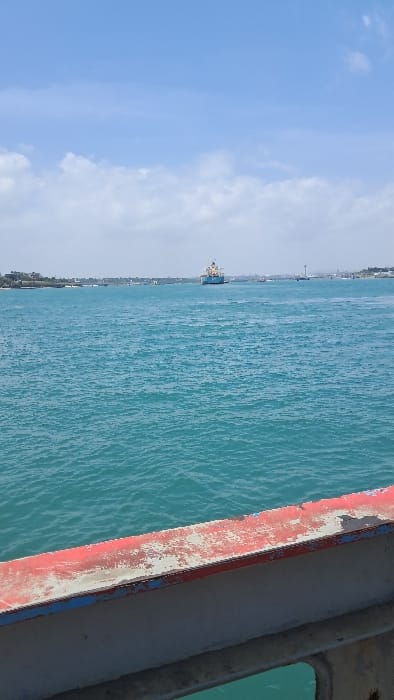 The image displays a wide view across a body of water identified as part of the Mombasa Old Port waterway, likely from the Likoni Ferry channel or a similar vantage point within the Ganjoni/Shimanzi ward, Mombasa, Kenya. In the immediate foreground, a weathered metal structure, possibly a railing or deck edge, occupies the lower section of the frame. This structure exhibits a prominent horizontal band of faded, peeling red paint over a grey metallic base, revealing rust and lighter underlayers, with a thin blue stripe below. The midground is dominated by an expanse of turquoise-blue water characterized by numerous surface ripples. In the distant background, a large blue and white cargo ship is positioned centrally on the water. Distant shorelines are discernible on both the left and right, featuring sparse vegetation and indistinct low-lying structures. The sky above is light blue with scattered white cumulus clouds. No individuals, explicit actions, or direct interactions are identifiable within the frame.