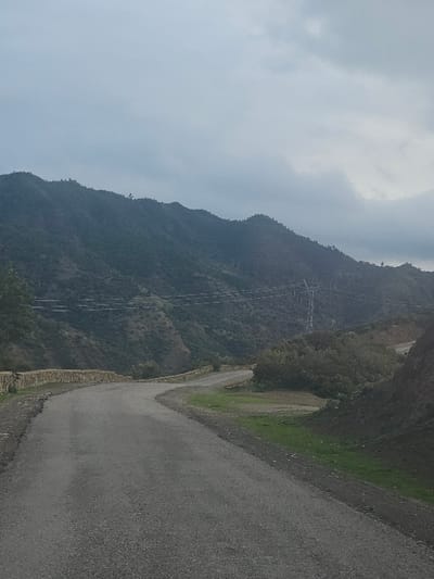 A winding asphalt road curves along a mountain slope. Lush green vegetation flanks the road on one side, contrasting with the steep, rugged terrain. Utility lines stretch across the mountain. The sky is overcast, creating a muted, somewhat hazy atmosphere. The road's edge is lined with a low stone wall.