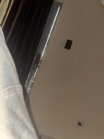 An indoor scene in Sabon Gari South, Nigeria, is depicted from a low angle. The upper left quadrant shows a section of a light-colored, possibly faded blue or grey, fabric sleeve belonging to an unseen individual. To the right of the sleeve, a dark door or partition is visible, with a narrow, vertical sliver of bright light emanating from a gap between its edge and the frame. A silver-toned door handle with a lock mechanism is present on the dark door. The wall occupying the central and right portions of the image is light beige. Mounted on this wall, in the upper right, is a black rectangular electrical component, possibly a switch or small device, positioned above a white switch plate. In the lower right, a white electrical wall outlet is present, with a black plug inserted into its lower receptacle, from which a thin white cable extends downwards. The room's ambient lighting appears dim, with the primary illumination originating from the bright gap at the door.