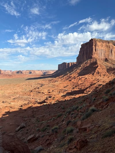 The image depicts a vast desert landscape in Oljato-Monument Valley, United States, under a bright blue sky with scattered white clouds. Dominating the right side of the frame is a massive, sheer red rock mesa, its face deeply grooved and casting a long shadow across the arid terrain. In the distance, a series of similar, smaller mesas stretch towards the horizon, forming a dramatic and rugged panorama. The foreground is a rocky, reddish-brown slope dotted with sparse, dry vegetation. The overall scene conveys a sense of isolation and the raw beauty of the desert Southwest.