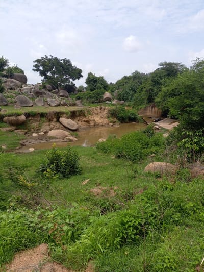 The image depicts a rural landscape in Rinjim Gani, Nigeria, characterized by a muddy stream or pond surrounded by lush vegetation and large rocks. A small, concrete structure, possibly a bridge or culvert, is visible spanning the water. The terrain is uneven, with eroded earth banks leading down to the water's edge. The sky is overcast with scattered clouds. The scene appears natural and undisturbed, with no human presence or activity immediately apparent. The time of day is indeterminate due to the diffused lighting conditions.
