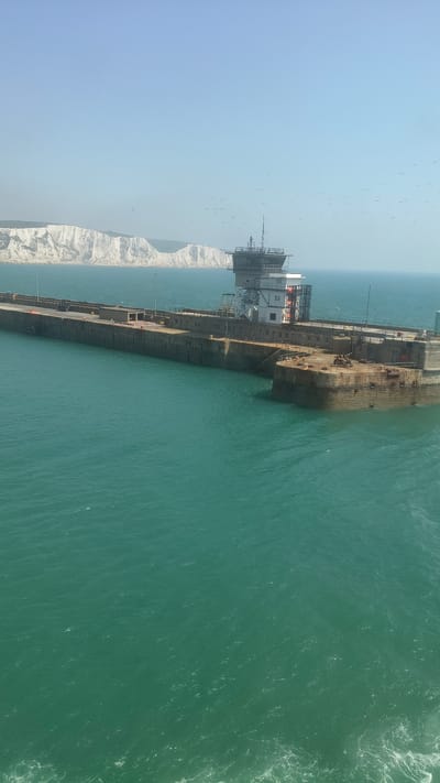 This is a high-angle, vertically oriented photograph showing a long, industrial-looking structure extending into a turquoise body of water. The structure appears to be a pier or a breakwater, with a weathered, rusty metal surface and several buildings or platforms built on top of it. One of these structures has white sections and appears to be a control room or administrative building. The water is choppy, with whitecaps suggesting some wind.
In the background, a white cliff face is visible, characteristic of the Dover coastline. The sky is a pale blue, indicating it's likely daytime, possibly morning or afternoon. Numerous small birds are scattered across the sky, some flying in flocks.
The overall scene depicts a functional, perhaps somewhat stark, maritime environment. There is no clear activity or event taking place, but the presence of the pier and the water suggests a port or coastal area. The lighting is bright, but there are no strong shadows to indicate a specific time of day. The location cues like the white cliffs strongly suggest Dover, United Kingdom. No text is visible in the image.