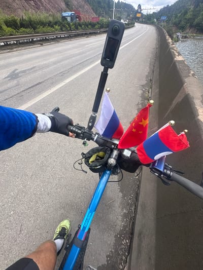 A first-person perspective shot shows a person riding a blue bicycle on a paved road with a concrete barrier on the right and a body of water beyond. The bicycle has multiple flags attached to its handlebars: the flags of Russia, China, and Laos. A black handlebar bag is attached to the handlebars. A digital display is mounted on the handlebars. A black 360-degree camera is mounted on a pole extending from the handlebars. The rider's left hand, wearing a blue sleeve and black glove, is on the handlebars. The rider's left leg, wearing a black cycling short and a neon green athletic shoe with black laces, is visible. The road appears to be a highway or a major road with multiple lanes and guardrails. In the background, there are hills covered with vegetation and some distant buildings. The sky is overcast.