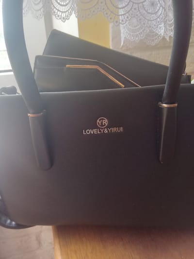This is a close-up, eye-level, indoor shot of a dark brown or black purse. The purse has a textured, leather-like finish. A logo is embossed on the front of the purse, which consists of a circle containing the letters "YR" in a stylized font, and the words "LOVELY & YIRUI" written below it in a smaller, silver font. The purse has two handles that are partially visible. In the background, a white lace trim is visible near the top of the frame, and a blurred yellow surface is behind that. A wooden surface is visible in the lower part of the frame. There are no people in the image. This image was taken in the city of Terebovlia, Ukraine.