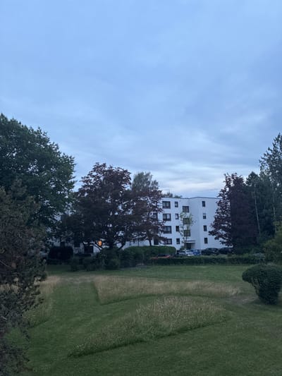 The image shows a residential building in the city of Memmingen, Germany. The building is white with a modern design and has several windows. In front of the building, there is a green lawn with some trees and bushes. A path is visible on the lawn, and there are cars parked in front of the building. The sky is cloudy and the light suggests it is either early morning or late evening.