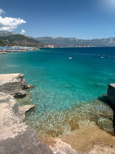The image depicts a panoramic view of a coastal scene in the city of Kubasi, Montenegro, under bright daylight. In the immediate foreground, the clear, shallow turquoise water reveals a rocky and sandy seabed with pronounced sunlight ripples. A light-colored concrete or natural rock formation is visible on the left, extending into the water, while a darker rock edge frames the right side.
The midground features deeper turquoise water, demarcated by a line of red and white buoys. Within this area, a person is seen on a white inflatable object, possibly a swan, and another individual is swimming. To the left, a populated beach area is visible, furnished with numerous blue sun umbrellas and lounge chairs, with multiple people present on the sand and in the shallow water. Buildings and lush green vegetation are situated on the hillside directly behind the beach.
The background presents a vast expanse of deeper blue sea, meeting a distant coastline characterized by a range of rugged, forested and rocky mountains. Scattered structures and settlements are discernible along this distant shoreline. A white sailboat is observed against the distant landmass on the right side of the frame. The sky is bright blue, featuring a few scattered white cumulus clouds, primarily clustered on the left side. The scene is illuminated by strong natural sunlight, indicative of a clear, sunny day.