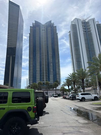 A bright green Jeep Wrangler is parked on a wet asphalt surface, with a large, dark-tinted spare tire mounted on its rear. The Jeep is positioned in the foreground, angled to the right, with its rear end facing the viewer. Behind the Jeep, a white SUV is parked further down the street. The scene appears to be a parking area or street beside a waterfront, as suggested by the presence of palm trees and reflections of buildings in the wet pavement.
In the background, three distinct skyscrapers dominate the skyline. The tallest building, a light-colored skyscraper with numerous balconies, is on the left. To its right, a darker skyscraper with blue-tinted windows and a tiered design stands tall. In front of these, a darker, more modern-looking skyscraper with a sloped top is visible. Palm trees are interspersed between the buildings and along the street, indicating a warm, tropical climate. The sky is partly cloudy with some wispy clouds and bright sunlight, suggesting it is daytime. The wet pavement implies recent rain or a high humidity environment. There are no people visible in the frame, and the overall mood is serene and urban, with a touch of outdoor adventure suggested by the Jeep. The word "RIMAMER" is visible on the rear window of the Jeep, written in reverse.