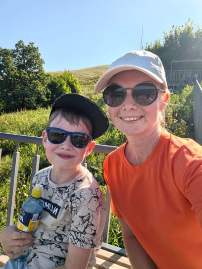 The image shows a selfie taken by a woman and a boy on a bench outdoors. The woman is wearing an orange t-shirt and a white baseball cap, with sunglasses. The boy is wearing a grey t-shirt with a black and white pattern and a black baseball cap, also with sunglasses. He is holding a bottle of water. They appear to be in a park or natural area, with green grass and trees in the background, under a clear blue sky. The location is specified as Akhmerovo, Russia.