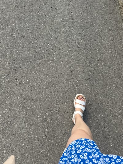 The image shows a person's legs and feet walking on a dark grey asphalt surface. The person is wearing white sandals and a blue dress with a white floral pattern. The scene is outdoors, with the asphalt surface likely a road or sidewalk. There is no visible indication of the specific location in Biczyce Dolne, Poland. The lighting suggests it is daytime.