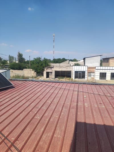 The image depicts an outdoor scene under a clear blue sky with a few scattered clouds. The foreground is dominated by a red corrugated metal roof, partially covered with dust and debris. A solar panel is visible on the left side of the roof, angled upwards. In the middle ground, a row of buildings is visible. Some appear to be in disrepair, with crumbling brick walls and open doorways. One building features a modern facade with large glass windows and white trim, suggesting it might be a shop or commercial space. Another building is constructed of light-colored bricks. Further in the background, trees and vegetation form a green backdrop, and a tall telecommunications tower stands prominently against the sky. The overall impression is of a somewhat dilapidated urban or semi-urban environment on a sunny day. The lighting suggests it is daytime, likely late morning or early afternoon, given the clear sky and the shadows cast on the roof. There is no visible text in the image.