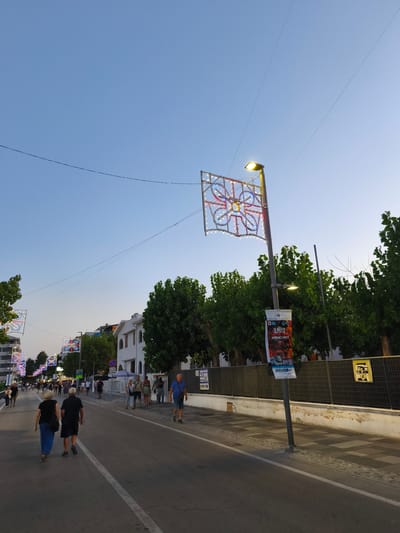 The image depicts a street scene in the city of San Salvo, Italy, during the early evening. The sky is clear and blue, transitioning to a lighter hue near the horizon. A street lined with trees and buildings is visible, with decorative lights strung across the street and illuminated on lamp posts. Several people are walking along the sidewalk and the street. One couple is walking away from the camera in the foreground, and other individuals and small groups are scattered further down the street, some appearing to be engaged in conversation or browsing.
Prominently featured are lamp posts adorned with square, illuminated decorations, each with a central circular motif and colorful lights. A banner hangs from one of these lamp posts, featuring text and images, possibly advertising an event. The buildings on the right are modern in style, with white facades. A black metal fence runs along the sidewalk on the right side of the street, behind which are trees. The street surface is asphalt with white lane markings. A sidewalk with a checkered paving pattern is visible to the right of the street. The overall atmosphere suggests a pleasant evening in a town or city, likely during a festival or summer gathering.