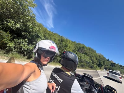 A woman and a man are riding a motorcycle on a road in Mišići, Montenegro. The woman is in the foreground, wearing a white helmet and a grey tank top. She is holding the camera, taking a selfie. The man is behind her, wearing a black helmet and a black and white vest with "EINTRACHT" printed on the back. They are on a sunny day, with green trees and a blue sky visible in the background. A white car is driving in front of them on the road.