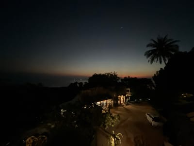 This is a nighttime aerial view of a coastal area in Karimunjawa, Indonesia, just after sunset. The sky transitions from a deep twilight blue at the top to faint hues of orange and pink near the horizon, suggesting the last remnants of daylight. A single bright star is visible in the upper right quadrant of the sky. In the foreground, a silhouetted palm tree stands against the dusky sky.
Below, a cluster of buildings with illuminated windows indicates a small settlement or resort. Lights are on inside the structures, and there appears to be some activity, with figures visible through the windows, possibly people dining or socializing. A dirt or gravel path winds through the scene, with a white car parked on it. The overall impression is one of a tranquil evening in a tropical location. The scene evokes a sense of peace and relaxation, characteristic of a vacation spot. There is no visible text in the image.