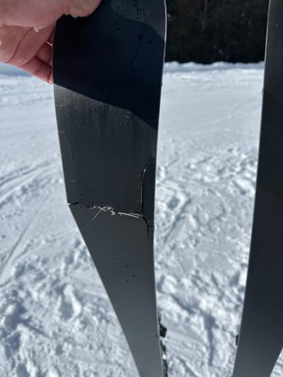 This image presents a close-up view of a fractured black ski held by a human hand, set against a snowy outdoor backdrop in Bad Hofgastein, Austria. The ski, held by what appears to be a bare or lightly-gloved hand visible at the upper left, displays a significant transverse fracture across its width, approximately one-third of the way down from the top. This break exposes white, fibrous composite material beneath the black top layer. Numerous smaller, linear cracks emanate from the main fracture, extending across the black surface and along the edges of the ski. A second, undamaged black ski is partially visible on the right side of the frame. The background consists of a snow-covered ground with discernible ski tracks, illuminated by direct sunlight, with a dark treeline visible in the distant background.