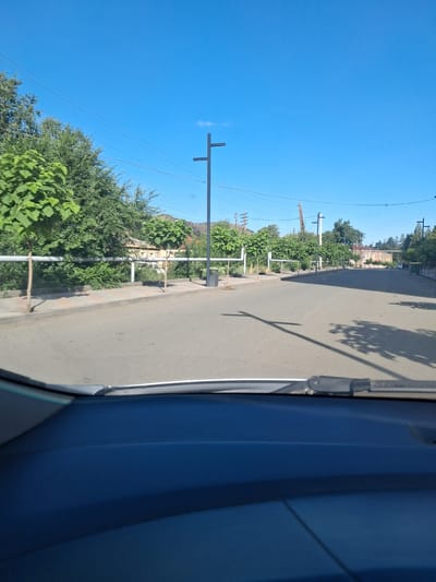 The image captures a street scene in Vayk, Armenia, under a clear blue sky. The perspective is from inside a vehicle, looking forward through the windshield. The street is paved and appears to be a two-lane road. On the left side of the street, there is a sidewalk with a row of trees. A white pipe runs along the edge of the sidewalk, supported by posts. Beyond the sidewalk and trees, buildings are visible, appearing to be residential or commercial structures. There are also more trees and vegetation in the background. On the right side of the street, the road continues into the distance, with shadows from trees cast on its surface. There are also some streetlights visible along the road. In the far background, hills can be seen. The overall impression is of a quiet, sunny day in a town or city.