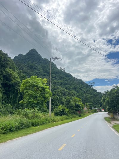 The image shows a paved road winding through a lush, mountainous landscape in Thuận Hòa Commune, Vietnam. The road is gray with yellow dashed lane markings. On the left side of the road, a steep, tree-covered mountain rises dramatically, its peak obscured by clouds. The vegetation is dense and vibrant green, with some vines climbing the mountainside. A tall utility pole with wires runs along the side of the road, casting shadows. On the right side of the road, more trees and foliage are visible, along with a few buildings and signs in the distance. The sky is partly cloudy with patches of blue visible. The overall impression is one of natural beauty and rural tranquility.