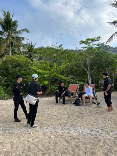 In Đông Ninh Hòa, Vietnam, on a sandy beach dotted with dried coral fragments and small pink flowers, a group of individuals are gathered amidst lush green trees and palm trees. Two people stand on the left, one holding a camera with a gimbal. In the center, two men are seated on orange lounge chairs, one in a blue shirt and white shorts, the other in a black t-shirt. To the right, another man stands, observing the scene. Beach chairs, bags, and footwear are scattered on the sand, suggesting a relaxed atmosphere. The sky above is a blend of blue and white clouds, with the indistinct shape of mountains visible in the background.