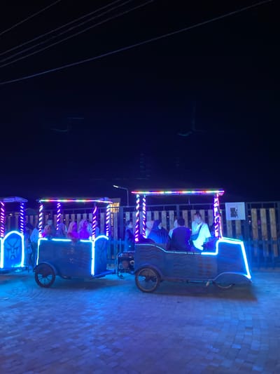 In Pardomuan I, Indonesia, two decorative, open-air vehicles designed to resemble train cars are illuminated by vibrant blue and rainbow-colored LED lights at night. The vehicles, which appear to be a form of amusement ride, are carrying passengers. The vehicle on the left has intricate blue lighting patterns along its sides and a prominent stylized mask-like design on its front. The vehicle on the right is also adorned with colorful lights, particularly along its roofline and edges. Several individuals, some wearing headscarves, are seated within the passenger compartments of both vehicles, suggesting they are enjoying a leisurely ride. The ground beneath the vehicles is paved with interlocking stones, which are also reflecting the blue light. A wooden fence lines the background, and the overall atmosphere is dark, indicating nighttime. The presence of power lines and what appears to be a light pole in the distance suggest an urban or semi-urban setting. The scene captures a moment of nighttime entertainment.