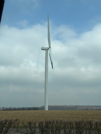 The image presents a rural landscape in Pagny-sur-Meuse, France, dominated by a large wind turbine under a partially clouded sky. A single, tall, slender white wind turbine shaft stands centrally, topped with a gray nacelle. Two of its three blades are visible; one extends vertically upwards, while the other angles downwards to the right. The blades appear stationary. In the immediate foreground, a dense, dark brown, leafless hedge occupies the lower portion of the frame, with glimpses of a dark grey surface, possibly a road, visible through its branches. Beyond the hedge, a vast, flat agricultural field covered in light brown stubble or harvested crops stretches across the midground. A dark green-brown treeline or forest forms the distant horizon. The sky features patches of bright blue with distinct white cumulus clouds on the left, transitioning to a more overcast, grayish cloud cover towards the right. Multiple light reflections and faint rectangular glares are visible across the image, suggesting it was captured through a window.