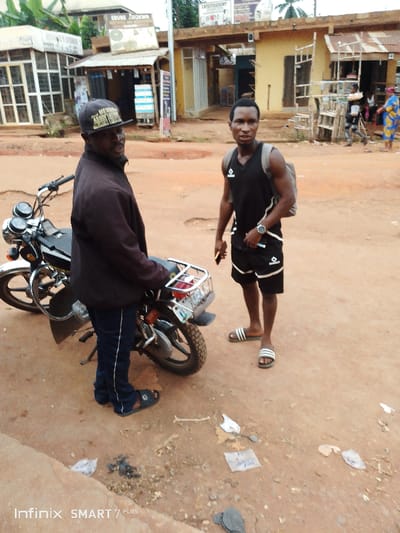 Two men stand on a dirt road, with a black motorcycle beside the man on the left. The man on the left wears a dark jacket, blue pants, a black cap, and sandals. The man on the right wears a black athletic shirt and shorts with white stripes, a watch, sandals, and a backpack. Buildings line the background, and trash litters the ground. An "Infinix SMART 7 PLUS" watermark is at the bottom.