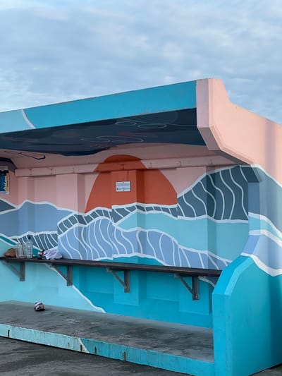 The image depicts an outdoor seating area with a mural on the back wall. The mural features a stylized depiction of waves and a setting sun in pastel colors, with shades of blue, pink, and orange dominating the scene. The structure itself is made of concrete, painted in bright blue and pink. A dark wooden bench is mounted on the back wall, with metal brackets supporting it. On the bench, there are several items: two plastic water bottles, a crumpled white cloth or bag, and a brown paper bag. In front of the bench, on the concrete ground, a single sandal is visible. The overall environment appears to be a public space, possibly a rest stop or a viewing area, with a cloudy sky overhead. The location is identified as Terhillion, Ireland.