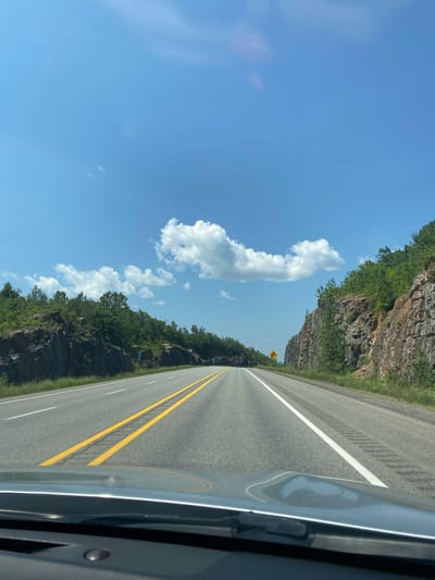 A first-person perspective shot captures a highway road with a bright blue sky and scattered clouds. On either side of the road, there are rocky cliffs with green trees growing on top and along the sides. The road is paved and has yellow and white lines marking the lanes. In the distance, the road curves to the right, disappearing from view. There is a yellow warning sign on the right side of the road in the distance, indicating a curve to the right. The foreground shows the dashboard of a car. There are no people or animals visible in the image. The scene appears to be a natural landscape with a man-made road.
