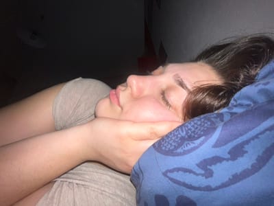 A person lies sideways with their head under a blue patterned blanket. Their eyes are closed, and their face is turned towards the viewer. They have brown hair, fair skin, and a neutral expression. Their arm is bent, with their hand near their neck. They wear a gray top. The background is a mix of light wall and dark space.