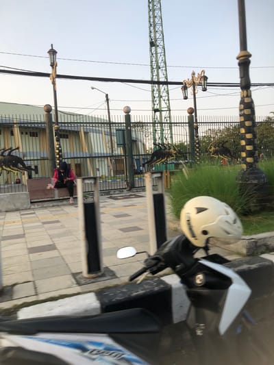 The image captures a street scene in Sidoarjo, East Java, with a focus on a motorcycle in the foreground. The motorcycle, equipped with a white helmet on its handlebars, is parked near a sidewalk. Beyond the motorcycle, a paved walkway with checkered tiling leads to a fence. A person wearing a pink jacket and a black hijab is seated on a bench behind the fence, looking at their phone. The background features a building with light green walls and a metal fence. Several decorative statues resembling prehistoric creatures are mounted on the fence. A tall, green lattice tower stands in the distance, suggesting an urban or park setting. Decorative streetlights are also visible, one with a classic design and multiple lamps, and another more modern, simpler design. The sky is a pale blue, indicating it is either early morning or late afternoon.