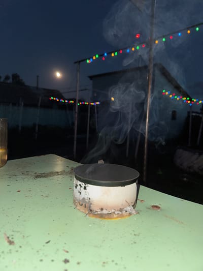 The image shows a close-up of a green metal surface with a pipe attached to it. Smoke is coming out of the pipe, suggesting an active process such as cooking or burning. In the background, a string of colorful lights, red, green, blue, and yellow, are visible, draped across a dark, outdoor setting. The sky is dark, indicating it is nighttime, and a bright, crescent moon is visible. The overall impression is of an outdoor gathering or event taking place at night, possibly a barbecue or a celebration, with a smoky atmosphere. The green surface appears weathered and possibly part of an outdoor appliance or structure. There is no visible text in the image. The context provided, Rodino, Russia, suggests a rural or semi-rural setting.