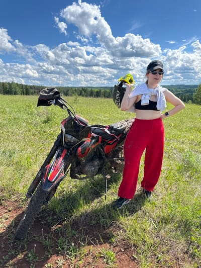 The image presents a young woman casually reclining on a parked, muddy dirt bike in a grassy field. She is wearing red sweatpants, a black crop top, sunglasses, and a baseball cap, with a white sweatshirt draped over her shoulders. She holds a yellow and black helmet in one hand and rests the other on her hip.
The dirt bike is black and red and appears well-used. The field is covered in green grass, and the ground around the bike is disturbed, revealing brown soil. In the background, a treeline borders the field under a bright blue sky filled with fluffy white clouds. The city name Пепеляева, Russia does not correlate with any specific details found in the image.
