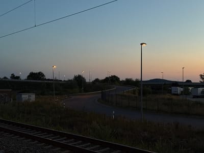 The image depicts a twilight scene on the outskirts of Forchheim, Germany. In the foreground, a set of railroad tracks runs diagonally from the lower left corner towards the center right. Beyond the tracks, tall, dry grass and vegetation line the landscape. A winding paved road is visible in the middle ground, illuminated by several streetlights. The road curves through an area with sparse trees and bushes. In the background, a low fence separates the road from a field with several parked white trailers or trucks, suggesting an industrial or commercial area. The sky transitions from a pale blue at the top to a warm orange and pink hue near the horizon, indicating either sunrise or sunset. Electrical wires stretch across the upper portion of the image. A small, grey, box-like structure with ventilation grilles is situated in the overgrown area to the left of the road. The overall atmosphere is calm and slightly deserted, characteristic of the early morning or late evening in a semi-industrial zone. No people are present in the image.