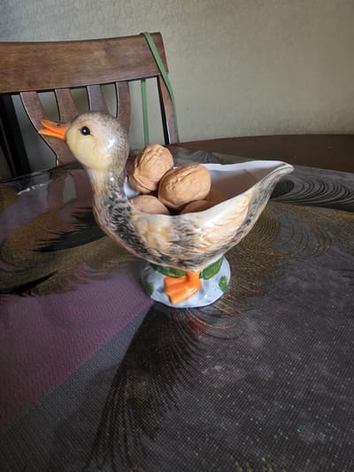 A ceramic duck figurine filled with walnuts sits on a table covered with a patterned tablecloth. The duck is positioned to the left of the frame, with its head turned to the left, beak open. The figurine has a base shaped like orange duck feet and green leaves. Behind the duck, a wooden chair with a green strap is visible. The background consists of a textured wall. The scene appears to be indoors. The image does not provide any information about the location of Levkovo, Russia.
