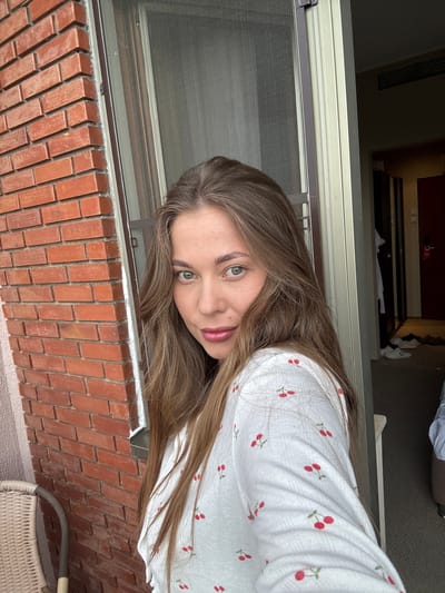 A young woman is seen from the chest up, positioned in front of a brick wall and a window with a screen. She has long, light brown hair and green eyes. She is wearing a white long-sleeved shirt with a cherry pattern. Behind her is a screen window, a brick wall, and what appears to be a doorway leading to an interior space with clothing visible. The bottom right corner shows a wicker chair. The location is likely a balcony or an area near the exterior of a building. It is impossible to ascertain the exact location in the city of Sosnovy, Russia without additional information or context.