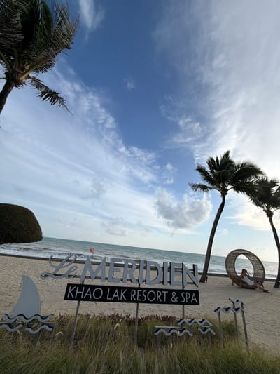 In Bang Muang, Thailand, a beach scene unfolds under a cloudy sky with visible palm trees. A sign with white lettering on a black background reads "KHAO LAK RESORT & SPA" below a larger, cursive "Le MERIDIEN." The beach features sand, gentle waves lapping the shore, and a red flag visible in the distance. To the right, a woman sits in a circular, woven chair, and two palm trees lean towards the ocean. In the foreground, decorative metal wave-like structures are partially obscured by tall, dry grass.