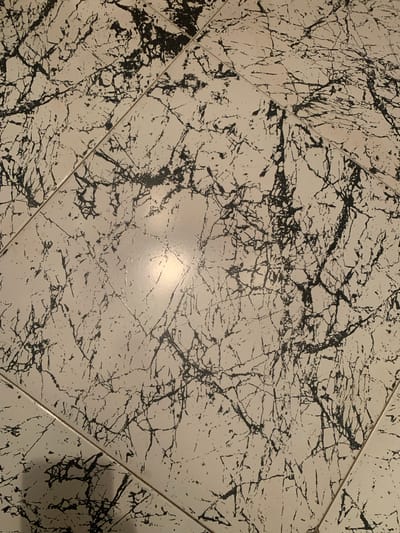 The image shows a close-up, overhead view of a tiled floor. The tiles are square and have a white or off-white base color with a black, vein-like pattern resembling marble or granite. The pattern is irregular and varies across each tile. There are visible grout lines separating the tiles, indicating a floor covering. A bright light source is reflecting off the glossy surface of some tiles, creating a shiny patch in the center-right portion of the image. There are no people or any specific objects clearly identifiable in the frame, nor any contextual clues suggesting a location in Tabagrebi, Georgia. The scene is purely focused on the texture and pattern of the flooring.