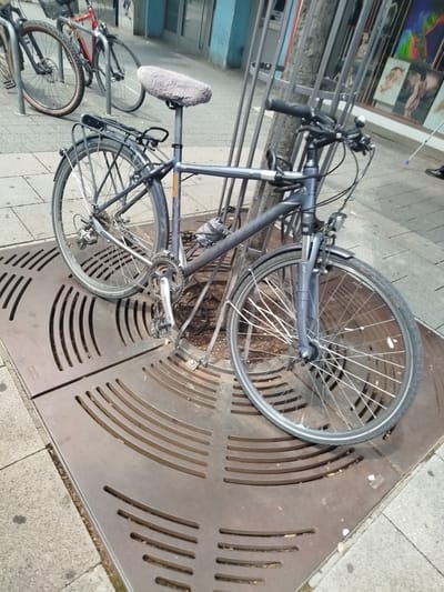 An upright, dark gray bicycle is parked on a grate in a city environment. The bicycle has a rear rack, a furry gray seat cover, and a bell on the handlebars. In the background, another bicycle with a red frame is parked. The scene appears to be a street or sidewalk, with buildings and a storefront visible in the distance. There are no people clearly visible in the image. The location is identified as Offenbach am Main, Germany.