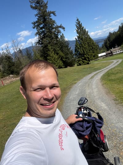 A man in a white T-shirt stands outside on a sunny day. He is holding a golf club. Behind him is a golf cart with golf clubs. Beyond the golf cart is a gravel path surrounded by green grass. Trees are on the side of the path. In the distance, there is water and mountains. The location is Bowen Island Municipality, Canada.