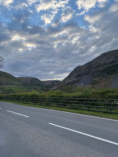 The image shows a road in Llangynog, United Kingdom, bordered by a metal safety fence and grass on one side and a mountain on the other. The asphalt road has white lane markings. A dramatic sky with textured clouds fills the top left of the frame.