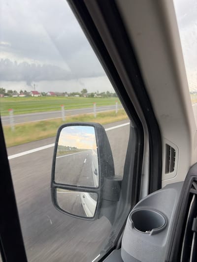 The image is a point-of-view shot from inside a vehicle, looking out the passenger side window and at the side-view mirror. The vehicle appears to be moving on a paved road, evidenced by the blurred scenery and road markings. In the foreground, on the right, is a gray interior panel with a cup holder and an air vent. The side-view mirror reflects a road scene with a white line marking the lane, and a white vehicle behind. Beyond the road, there is a green grassy field with red-topped posts marking its edge, and a row of houses with red roofs in the distance. The sky is overcast with dramatic cloud formations. The location is identified as within the Станькаўскі сельскі Савет, Belarus, suggesting a rural or semi-rural setting.