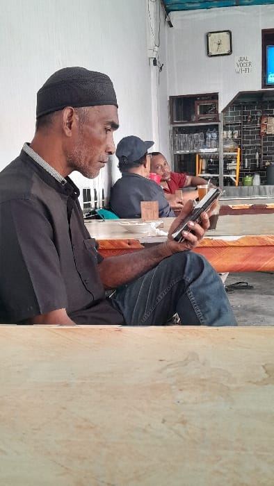 Three men are sitting down, seemingly on a ledge. The man closest to the camera is wearing a black cap and shirt. The man in the middle is wearing a black baseball cap and grey shirt. The man furthest away is wearing a red shirt. The man closest to the camera is holding a cell phone. The background is a white wall with a window. There is also a clock and text on the wall.