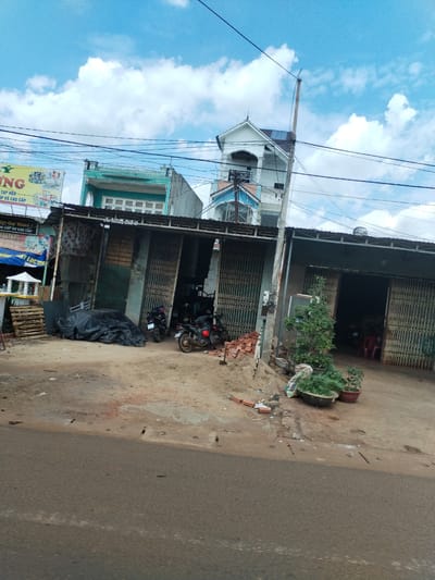 The image shows a street scene in Thôn 4, Vietnam, with a row of buildings on the right side of the road. The buildings have metal roller doors, some of which are closed. Two motorcycles are parked in front of one of the buildings. There are potted plants near the entrance to the buildings. A pile of bricks is visible on the ground near the buildings. The road in the foreground is paved. A sign above the buildings says "ING TẠP HÒA, CẤP VÀ CAO CẤP". Overhead, there are numerous power lines. The sky is blue with scattered clouds.