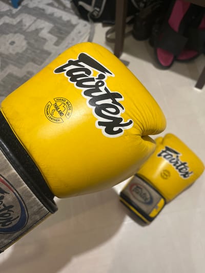A pair of yellow Fairtex boxing gloves is depicted in an indoor setting. The specific location in Clermont-Ferrand, France, is not identifiable from the visual information. The foreground features one prominent, slightly worn yellow glove with black 'Fairtex' branding. A circular stamp on this glove indicates 'HAND MADE IN THAILAND' and 'GENUINE LEATHER' with a map of Thailand. The wrist strap is a grey, white, and blue material with additional Fairtex branding. A second, identical yellow Fairtex glove rests out of focus in the background. The gloves are placed on a light-colored floor or surface. The background shows blurred elements including a patterned grey and white rug, the dark leg of a piece of furniture, and a pair of pink and black slide sandals underneath it. No people or active interactions are visible.