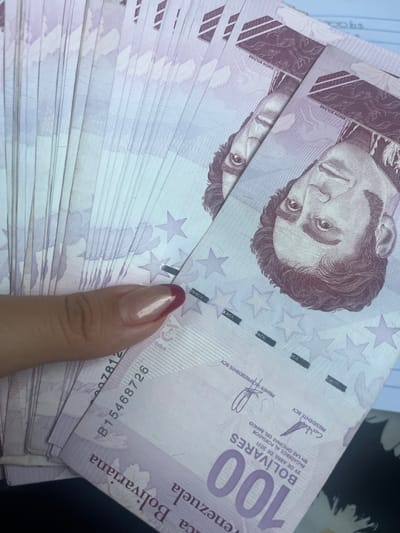A close-up, overhead shot displays a fan of Venezuelan 100 Bolivares banknotes. The denomination "100 BOLIVARES" is prominently visible on the lower right side of the bills. The portrait of Simón Bolívar appears on the upper right portion of the notes. A woman's hand, with a pinkish-gold colored nail polish and a dark red accent on the tip of one nail, is holding the banknotes. The background is blurred, but appears to be a flat surface. There is no indication of the specific location within Conuco Viejo, Venezuela.