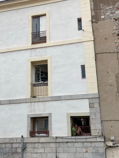 This image depicts a multi-story building facade in Sète, France, adjoined by an older, less maintained structure on the right. The primary building is finished in white stucco with cream-colored trim around windows and between floors, and features a grey stone block base.
On the top floor, a window with a small, rust-colored wrought-iron balcony is present on the left. A narrow, vertical window is visible to the right, on the building's side.
The middle floor features a large window with a rust-colored wrought-iron balcony. A black and white cat stands on the balcony ledge, looking outward. Inside the room, shelves are visible, and a green broom leans against the railing. A corresponding narrow, vertical window is on this floor.
On the bottom floor, a window with partially open wooden shutters and a decorative, dark red metal railing is on the left. To the right, a person in a green t-shirt and light-colored shorts sits on the balcony floor, leaning against the wall, holding a phone to their ear. A grey drainpipe descends the left side of the facade.
The adjacent building on the right exposes rough stone and patched concrete, traversed by multiple black electrical conduits and cables.